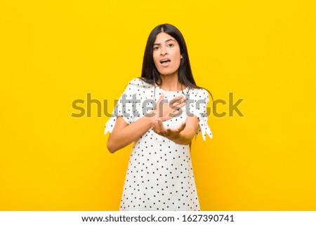 young pretty latin woman feeling happy and in love, smiling with one hand next to heart and the other stretched up front against flat wall