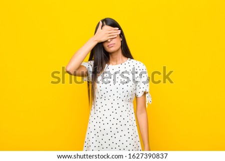 young pretty latin woman covering eyes with one hand feeling scared or anxious, wondering or blindly waiting for a surprise against flat wall
