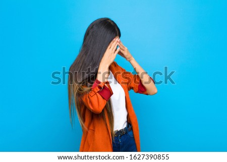 young pretty latin woman covering eyes with hands with a sad, frustrated look of despair, crying, side view against flat wall