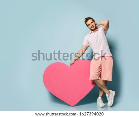 Young handsome man in white polo shirt hold pink heart shape toy present gift in hand on pastel blue wall background. Love gift for valentines day with text space