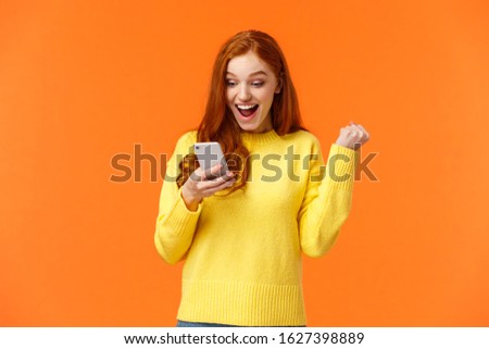 Girl received thousand likes under new picture. Cheerful and triumphing pretty redhead female fist pump say yes, cheering or celebrating good news, looking smartphone screen, orange background