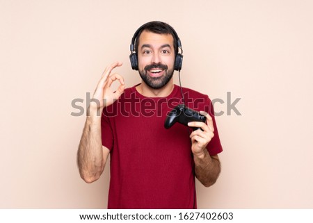 Man playing with a video game controller over isolated wall surprised and showing ok sign