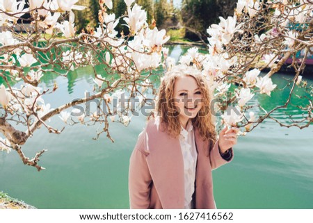 blonde girl in a pink coat stands by a blossoming magnolia on a background of green water and laughs merrily