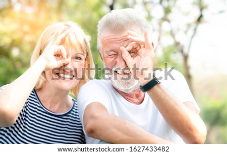 Happy grey-haired old man wearing T-shirt and senior woman wearing striped T-shirt make selfie in the park with happy face smiling doing ok sign with hand on eye looking through fingers.Body language.