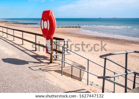 Life belt by the steps leading to a beach from a fenced path on a sunny spring day. An offshore wind farm is visible in background.