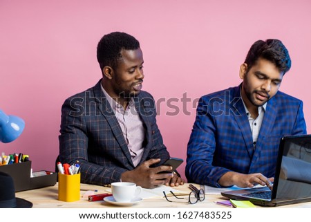 Shot of two businessmen wearing suits sitting in office at desk, using laptop and cellphone. Indian man sharing ideas with african american colleague, showing presentation of new project on computer.