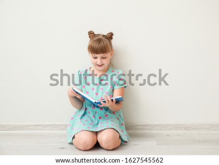 Cute little girl with book sitting near white wall