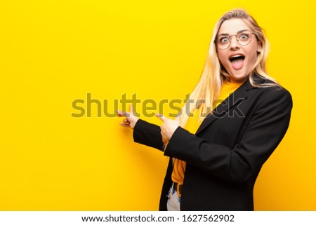 young pretty blonde woman feeling shocked and surprised, pointing to copy space on the side with amazed, open-mouthed look against yellow wall