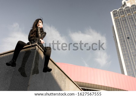 Girl all in black is citing on top of some architecture object, there are red wall and a skyscraper behind her. Fashion photo with gothic styled girl. A concept of a subculture, protest, mystique