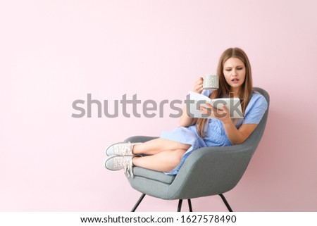 Beautiful young woman reading book and drinking tea while sitting in armchair near color wall
