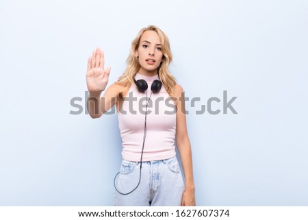 young blonde woman looking serious, stern, displeased and angry showing open palm making stop gesture listening music