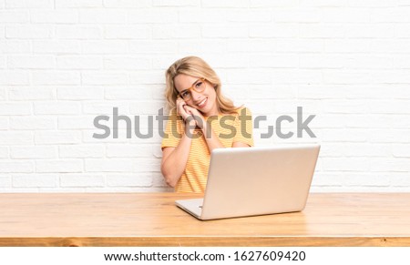 young blonde woman feeling in love and looking cute, adorable and happy, smiling romantically with hands next to face using a laptop