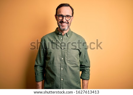 Middle age hoary man wearing casual green shirt and glasses over isolated yellow background with a happy and cool smile on face. Lucky person.