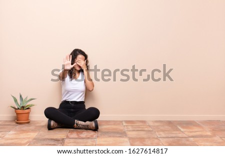young pretty woman covering face with hand and putting other hand up front to stop camera, refusing photos or pictures sitting a terrace floor