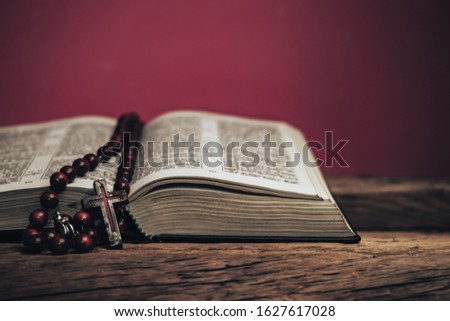 Open Holy Bible and beads crucifix on a old oak wooden table. Beautiful red wall background.