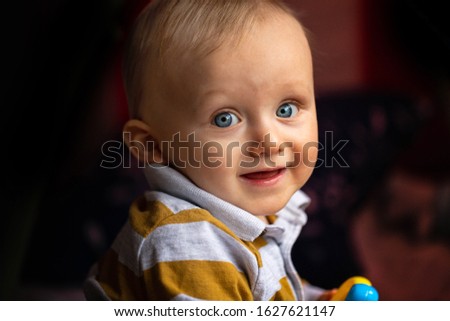 Smiling cute blond bue eyed baby boy model holding a toy wearing a grey mustard shirt on the dark blurry background. Artistic portrait close up with shallow depth of field. Adorable side posing