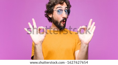 young crazy cool man feeling shocked, amazed and surprised, showing approval making okay sign with both hands against flat wall