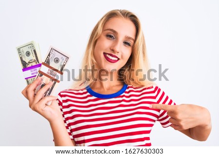 Beautiful woman wearing red striped t-shirt holding dollars over isolated white background with surprise face pointing finger to himself