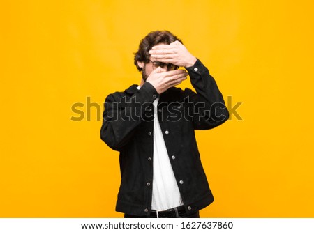 young crazy handsome man covering face with both hands saying no to the camera! refusing pictures or forbidding photos against orange wall
