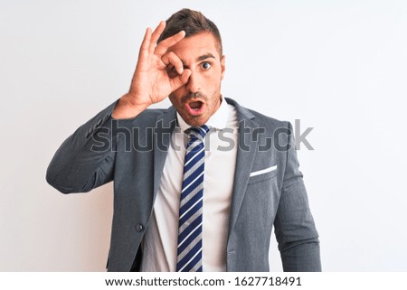 Young handsome business man wearing suit and tie over isolated background doing ok gesture shocked with surprised face, eye looking through fingers. Unbelieving expression.