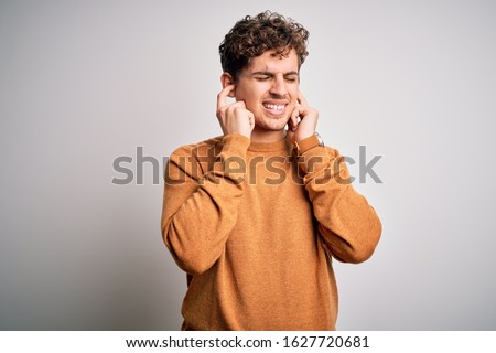 Young blond handsome man with curly hair wearing casual sweater over white background covering ears with fingers with annoyed expression for the noise of loud music. Deaf concept.