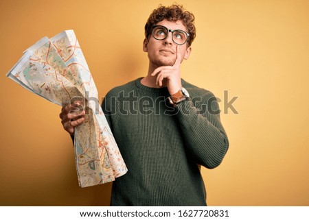 Young blond tourist man on vacation with curly hair holding city map over yellow background serious face thinking about question, very confused idea