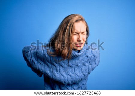 Young beautiful blonde woman wearing casual turtleneck sweater over blue background Suffering of backache, touching back with hand, muscular pain