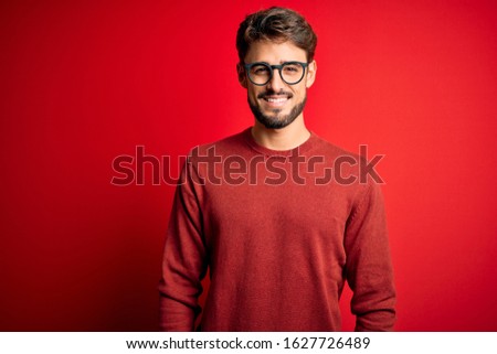 Young handsome man with beard wearing glasses and sweater standing over red background with a happy and cool smile on face. Lucky person.