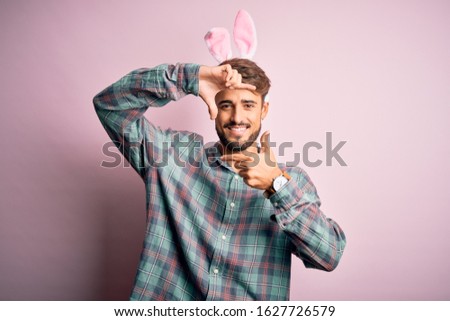 Young handsome adult man wearing cute easter rabbit ears over pink isolated background smiling making frame with hands and fingers with happy face. Creativity and photography concept.