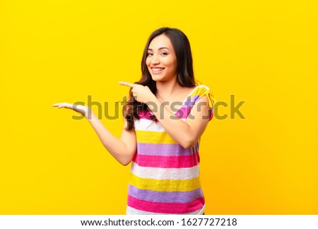 young latin pretty woman smiling cheerfully and pointing to copy space on palm on the side, showing or advertising an object against flat wall