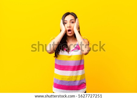 young latin pretty woman looking unpleasantly shocked, scared or worried, mouth wide open and covering both ears with hands against flat wall