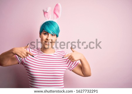 Young woman with fashion blue hair wearing easter rabbit ears over pink background looking confident with smile on face, pointing oneself with fingers proud and happy.