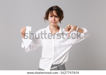 Dissatisfied young business woman in white shirt posing isolated on grey wall background. Achievement career wealth business concept. Mock up copy space. Hold down value fall arrow showing thumb down