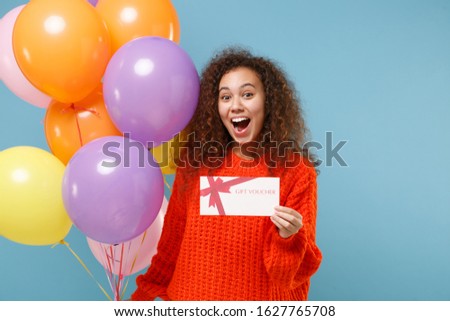 Excited young african american girl in casual orange knitted clothes isolated on pastel blue wall background. Birthday holiday party concept. Celebrating holding colorful air balloon gift certificate