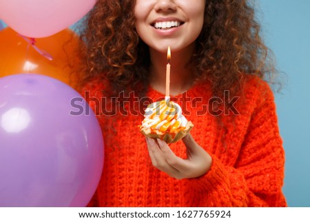 Cropped image of cute young african american girl in orange knitted clothes isolated on pastel blue background. Birthday holiday party concept. Celebrating hold colorful air balloons cake with candle