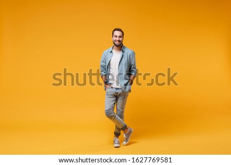 Smiling young bearded man in casual blue shirt posing isolated on yellow orange background, studio portrait. People sincere emotions lifestyle concept. Mock up copy space. Holding hands in pockets Royalty-Free Stock Photo #1627769581