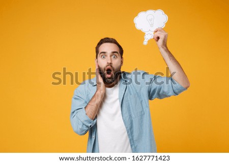 Shocked young man in casual blue shirt posing isolated on yellow orange background. People lifestyle concept. Mock up copy space. Hold say cloud with lightbulb, put hand on cheek, keeping mouth open