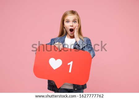 Shocked little kid girl 12-13 years old in denim jacket isolated on pink background. Childhood lifestyle concept. Mock up copy space. Hold huge like sign social network heart form, put hand on cheek