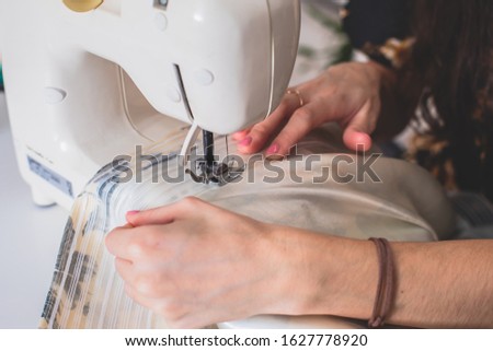 Process of sewing the curtains at home, close up of curtain tape on the sewing machine, hemming, tailoring, repairing and stitching cloth and dress, with the hand of female dressmaker in background
