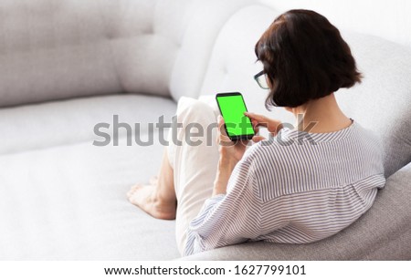 Middle-aged brunette woman with glasses on the gray sofa use green screen smartphone
