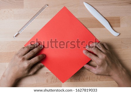 Top shot of both hand doing origami fold with a square red paper with bone folder and ruler besides