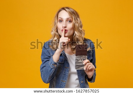 Secret girl in denim clothes isolated on orange background. Proper nutrition or sweet dessert dieting concept. Mock up copy space Hold chocolate bar say hush be quiet with finger on lips shhh gesture Royalty-Free Stock Photo #1627857025