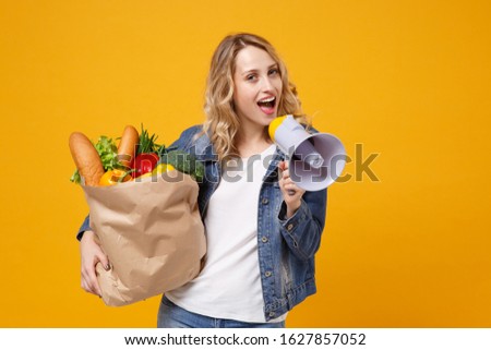Funny woman in denim clothes isolated on orange background. Delivery service from shop or restaurant concept. Hold brown craft paper bag for takeaway mock up with food products scream in megaphone