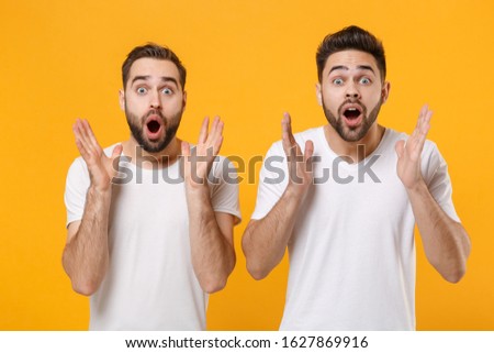 Shocked young men guys friends in white blank empty t-shirts posing isolated on yellow orange background in studio. People lifestyle concept. Mock up copy space. Keeping mouth open, spreading hands