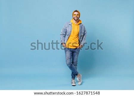 Smiling young hipster guy in fashion jeans denim clothes posing isolated on pastel blue background studio portrait. People emotions lifestyle concept. Mock up copy space. Holding hands in pockets Royalty-Free Stock Photo #1627871548