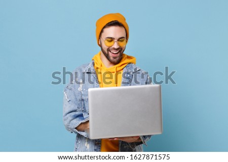Funny young hipster guy in fashion jeans denim clothes posing isolated on pastel blue background studio portrait. People emotions lifestyle concept. Mock up copy space. Working on laptop pc computer