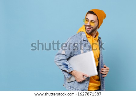 Side view of young hipster guy in fashion jeans denim clothes posing isolated on pastel blue background in studio. People lifestyle concept. Mock up copy space. Hold laptop pc computer, looking back
