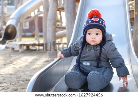 Cute baby boy playing in children’s slide at playground in the winter,he wearing warm clothes with hat. Happy mixed race Asian-German infant about 8-9 months old play outdoor park.