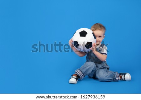 Сhild sits with a ball on a blue background and smiles, holding a soccer ball in front of him. Football competition. Sport concept of football, World Cup or European football tournament.
