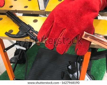 A welder man in red work gloves is working and holding an electron and clamps for welding at the table.